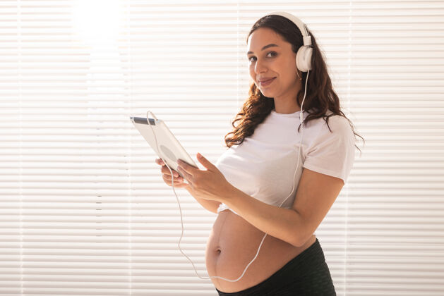 肖像美丽的年轻孕妇通过订阅选择音乐 并通过耳机收听音乐腹部预期