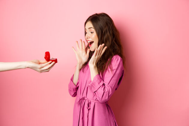 年轻惊讶的女友收到求婚的画像 看着手上拿着订婚戒指的人 惊叫着站在粉色的墙上迷人微笑积极