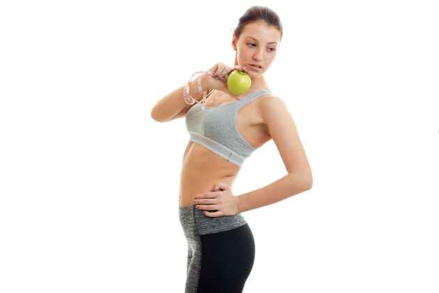 女士苗条的年轻女子侧身站着 低头 手里拿着隔离在白墙上的苹果锻炼健康磁带