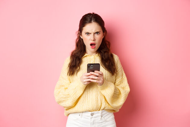 女性困惑和愤怒的女朋友在电话里读着奇怪的信息 盯着相机沮丧和质疑 站在粉红色的墙上兴奋应用程序时尚