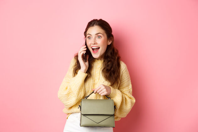 年轻快乐迷人的女孩在打电话的时候看起来很惊讶 在打电话的时候收到惊人的消息 拿着钱包 站在粉色的墙上成人女性休闲