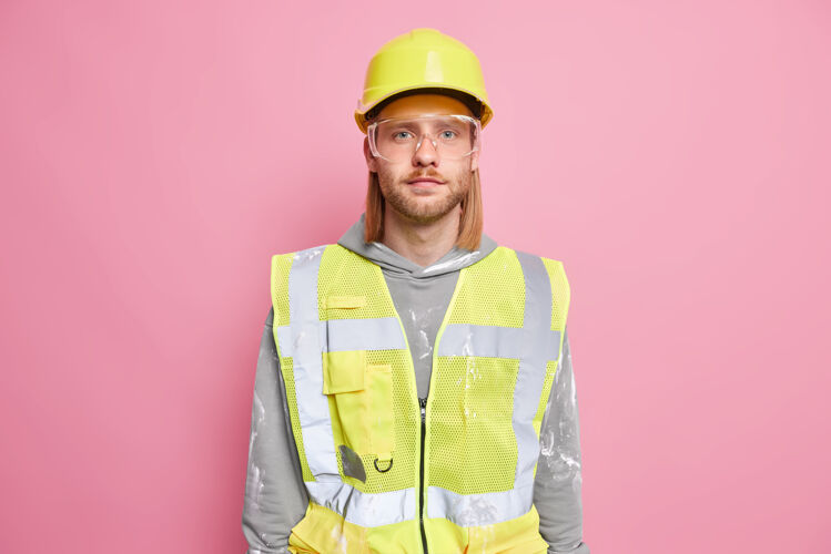 建筑师认真的建筑工人工程师戴施工安全帽制服眼镜看起来自信地准备工作隔离在粉红色的墙壁上自信的工人或建筑工人建筑工人头盔硬地