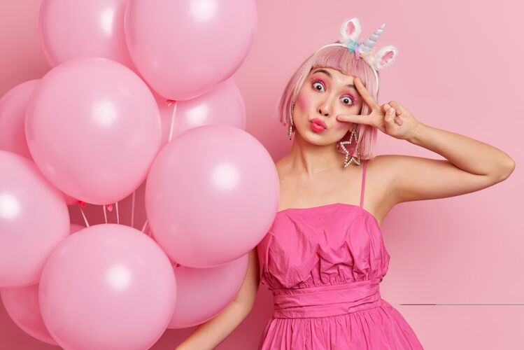 庆祝人们的生活方式庆祝节日的概念亚洲女人与鲍勃发型倾斜的头有有趣的表情手势胜利标志享受派对穿着时髦的礼服与气球的姿势独角兽粉色生日