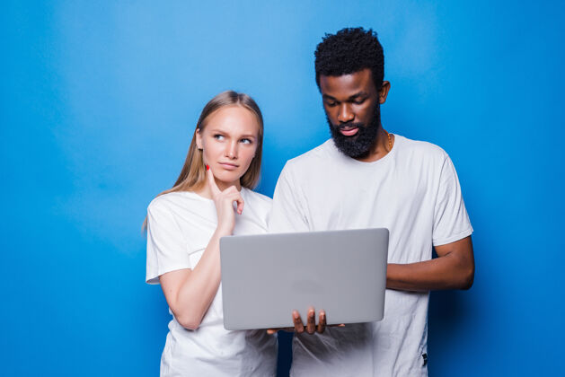 通信年轻的混血夫妇拿着笔记本电脑孤零零地站在蓝色的墙上人信使多样性
