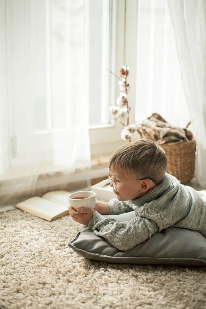 肖像一个穿着针织毛衣的漂亮小男孩拿着一杯热咖啡在窗边看书茶舒适秋天秋天的心情沙发聪明微笑
