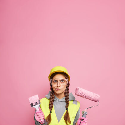 工人室内装修和家居装修严重不高兴的妇女穿着建筑服装 持有维修工具 重点放在上面准备油漆墙壁隔离粉红色墙壁空间颜色压路机设计师