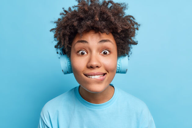 歌曲惊讶好奇的非裔美国妇女咬嘴唇的照片看起来直接戴着无线耳机穿着休闲t恤隔着蓝色的墙人们休闲的生活方式立体声女性积极