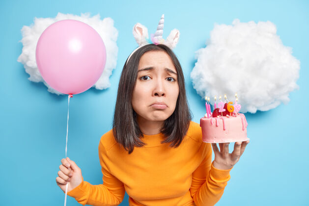 蛋糕深色头发的女人悲伤地看着相机独自庆祝生日 手里拿着美味的蛋糕和蓝色的充气气球生日女人黑发