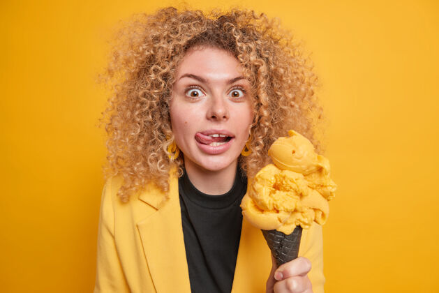 热量快乐惊喜的女人卷曲浓密的头发舔嘴唇伸出舌头拿着美味的冰淇淋诱惑着吃夏天的甜点摆在鲜艳的黄色墙壁上嗯 多好吃啊奇迹女人享受