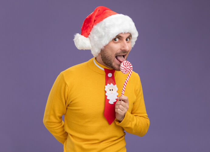 背部令人印象深刻的年轻白种人戴着圣诞帽打着领带拿着圣诞甜甜的手杖藏在后面看另一个舌头准备吃手杖隔离在紫色墙上显示男人隐藏