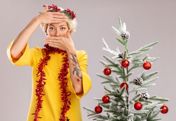 花环年轻的金发女人戴着圣诞花环 脖子上戴着金属丝花环 站在装饰好的圣诞树旁 双手放在额头和嘴巴上 隔离在白色的墙上近周围圣诞节