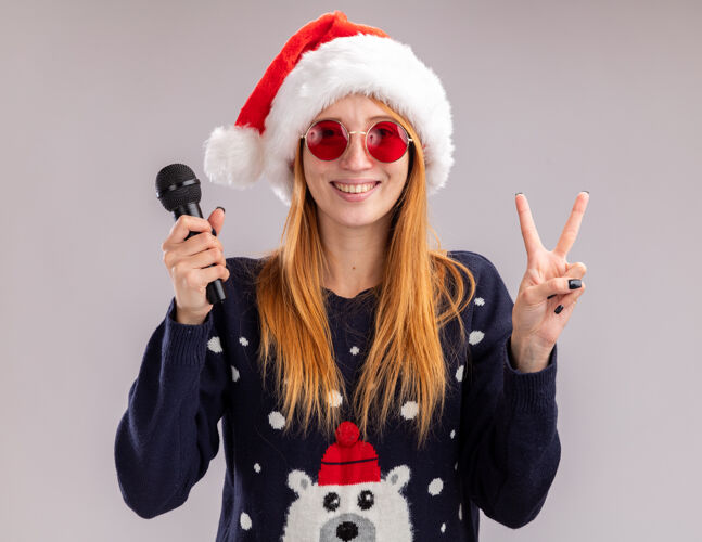 显示微笑的年轻漂亮女孩戴着圣诞帽 戴着眼镜 拿着麦克风 在白色的墙上显示出与世隔绝的和平姿态穿举行圣诞节