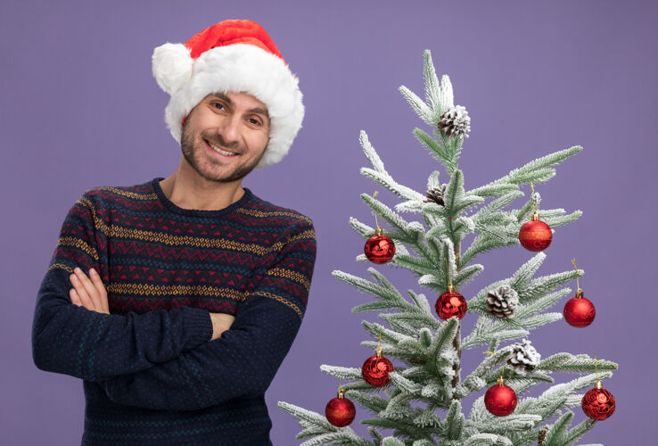 姿势带着圣诞帽微笑的白人年轻人站在装饰好的圣诞树旁 封闭的姿势 在紫色的墙上显得孤立无援年轻帽子近
