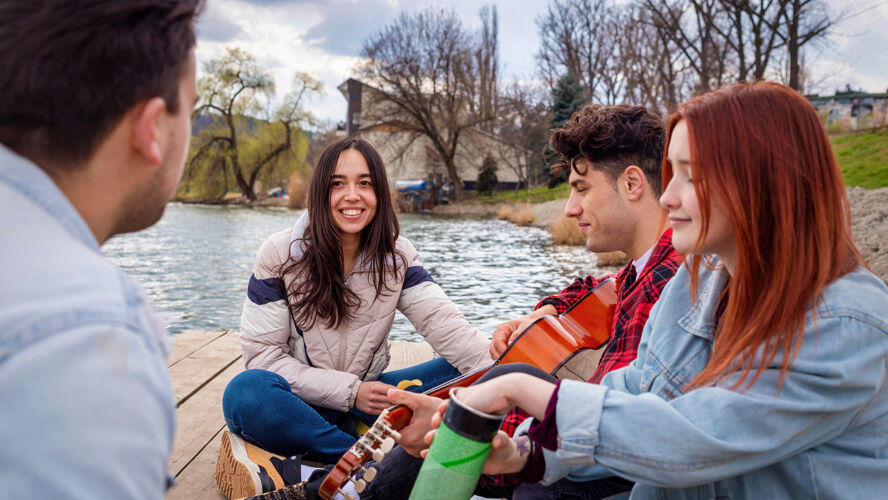 夏天四个年轻的朋友在公园的湖边唱歌 休息 弹吉他休闲花园自然