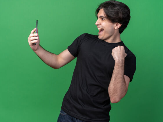 抱着快乐的年轻帅哥穿着黑色t恤 拿着手机 看着手机 在绿色的墙上显示“是”的手势穿着市民年轻人