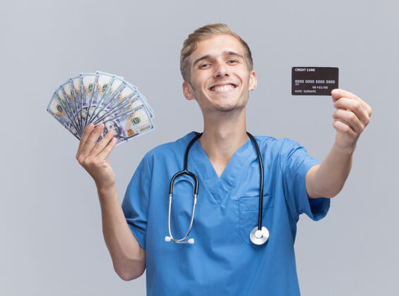 男性微笑的年轻男医生穿着医生制服 手持听诊器 拿着现金和信用卡对着隔离在白墙上的摄像机立场现金制服