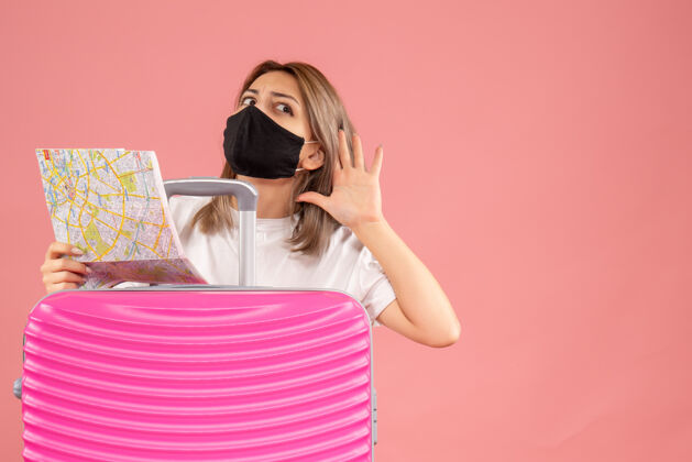 旅游正面图：戴着黑色面具的年轻女子拿着地图 听着粉色手提箱后面的东西漂亮成人冒险