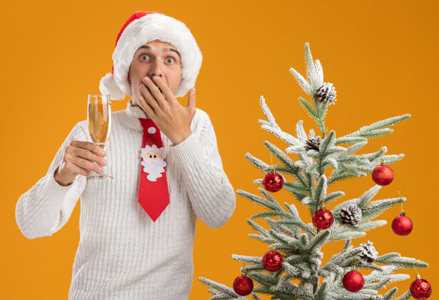 小伙子一个戴着圣诞帽 系着圣诞老人领带的年轻帅哥站在装饰好的圣诞树旁 手里拿着一杯香槟 手放在嘴边 在橙色的墙上显得孤立无援领带帅哥戴着