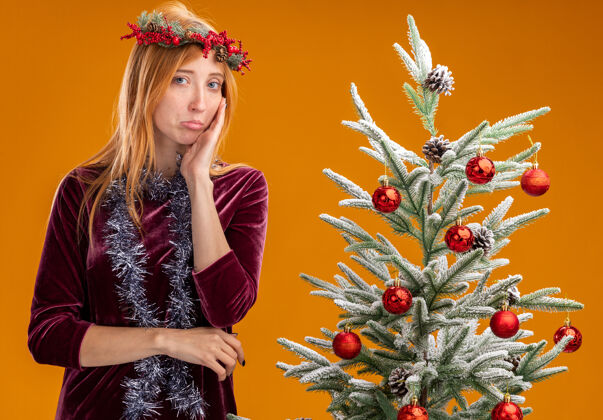 悲伤站在圣诞树旁的悲伤的年轻漂亮的女孩 穿着红色的裙子 脖子上戴着花环 手放在脸颊上 隔离在橙色的墙上花圈戴着在附近