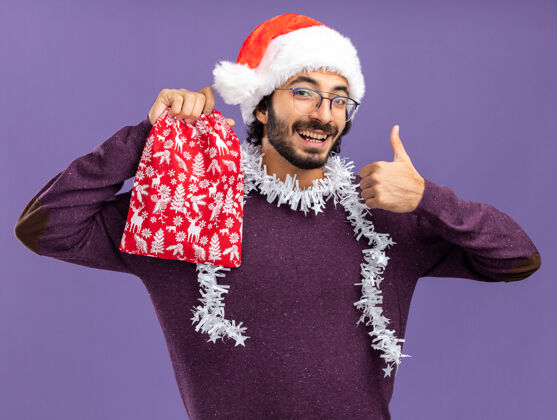 帅气微笑的年轻帅哥戴着圣诞帽 脖子上戴着花环 手里拿着圣诞包 在蓝色的墙上孤立地竖起大拇指抱年轻秀