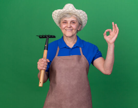老人带着微笑的上了年纪的女园丁戴着园艺帽拿着耙子 打着ok的手势绿色微笑园艺