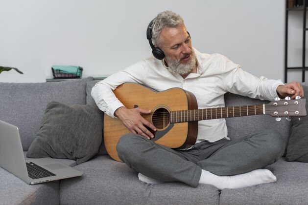 吉他课老人在家的沙发上用笔记本电脑学习吉他课胡须成人成熟