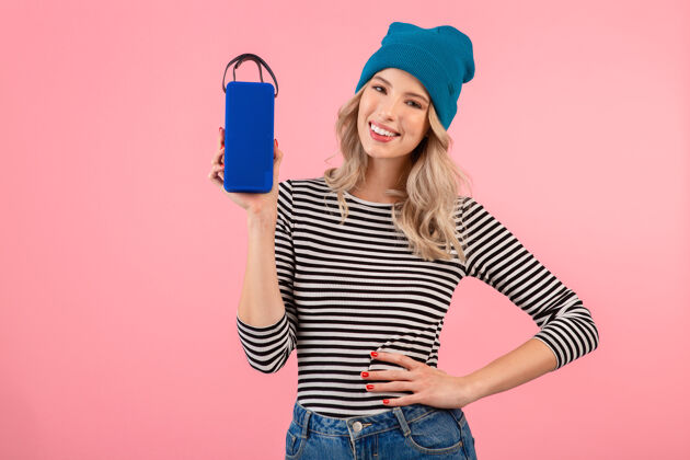 配件年轻漂亮的女人拿着无线扬声器听音乐穿着条纹衬衫和蓝色帽子微笑快乐积极的心情摆在粉红色的背景上科技无线服装