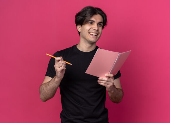 表情微笑着看着身边年轻帅哥穿着黑色t恤拿着笔记本用铅笔隔离在粉红色的墙上站着侧身笔记本