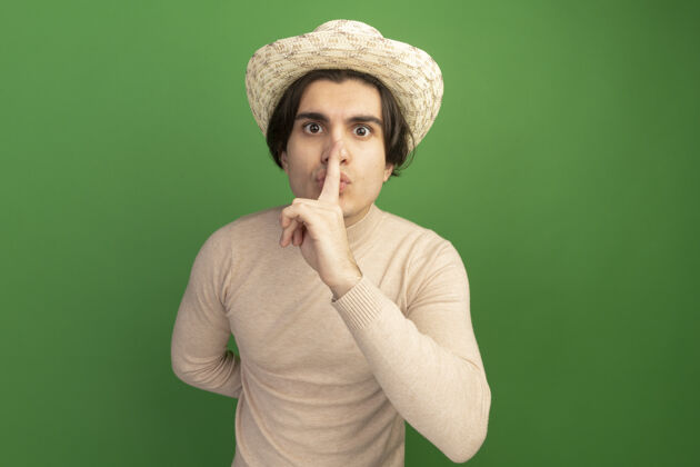 表演戴着帽子的年轻帅哥在绿墙上表现出沉默的姿态手势姿势男人