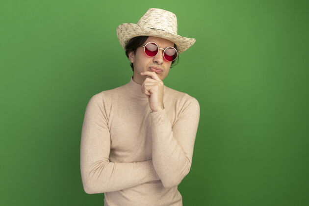 年轻自信的年轻帅哥戴着帽子 戴着眼镜 把下巴孤立地抓在绿色的墙上 留着临摹的空间表情人站着