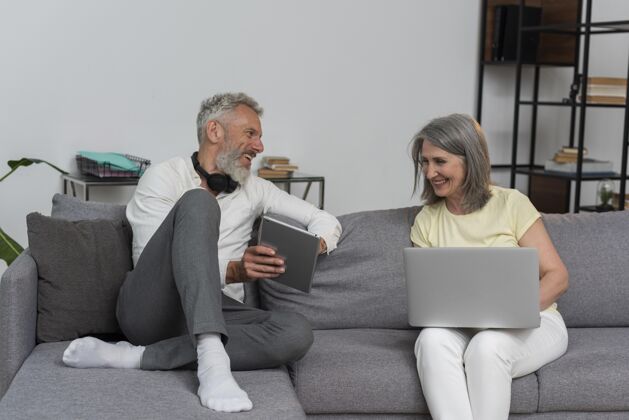 内部资深男女在家沙发上使用笔记本电脑和平板电脑设备老年人成人