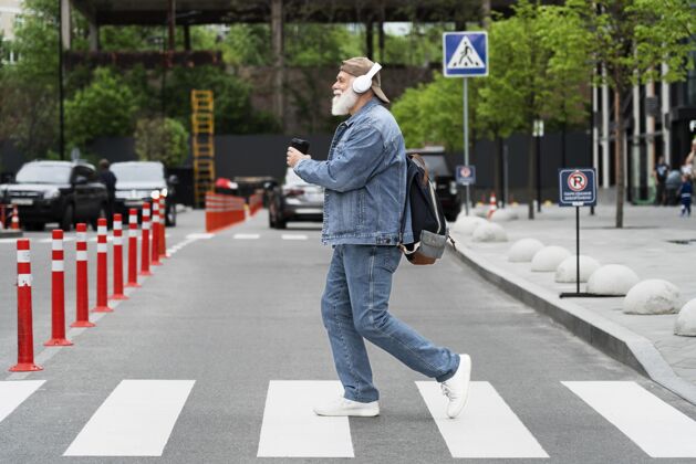 男人老人边过街边用耳机听音乐的侧视图退休老年人城市