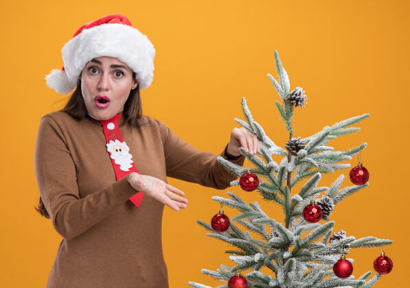 圣诞节惊讶的年轻漂亮女孩戴着圣诞帽打着领带站在旁边的圣诞树孤立在橙色的墙上穿站领带