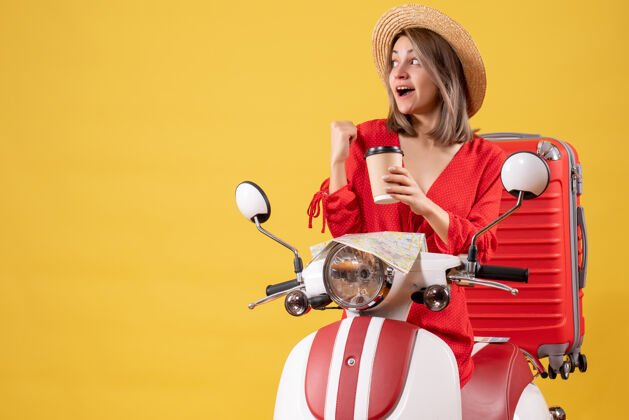 旅游正面图穿着红色连衣裙的年轻女士拿着咖啡杯指着后面靠近轻便摩托车的地方咖啡漂亮人