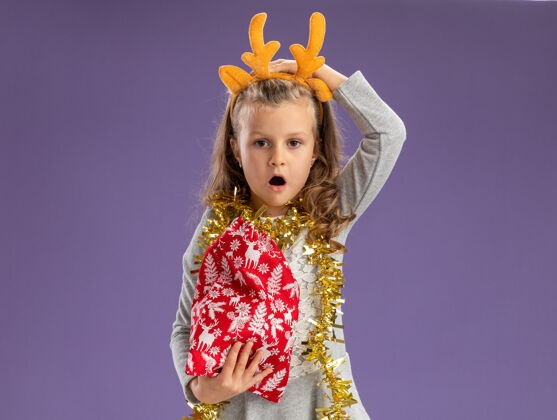 花环遗憾的小女孩戴着圣诞发箍 脖子上戴着花环 手里拿着圣诞包 手放在头上 孤零零地站在蓝色的墙上小圣诞节箍