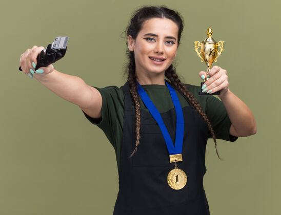 脸微笑的年轻女理发师身着制服 手持奖牌 手持优胜者杯 手持理发器 对着橄榄绿墙上孤立的镜头年轻人公民头发