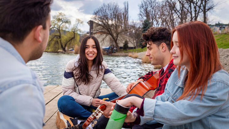夏天四个年轻的朋友在公园的湖边唱歌 休息 弹吉他休闲花园自然