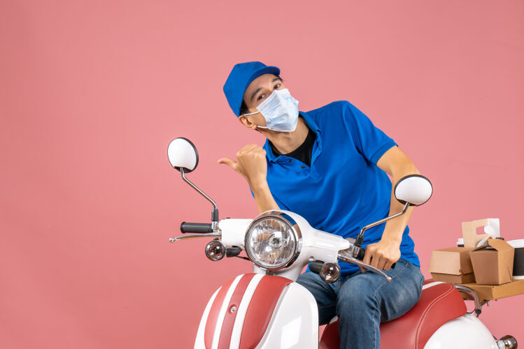 摩托车顶视图好奇的快递员戴着医学面具戴着帽子坐在滑板车上指着粉色背景上的东西穿桃医学