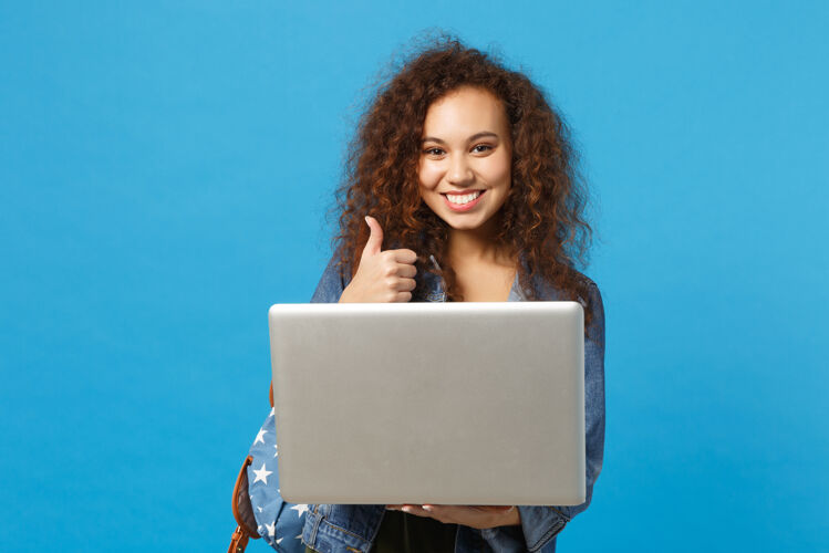 学生年轻的非洲裔美国女孩十几岁的学生在牛仔服装 背包工作的pc隔离在蓝色的墙壁上电脑打字持有