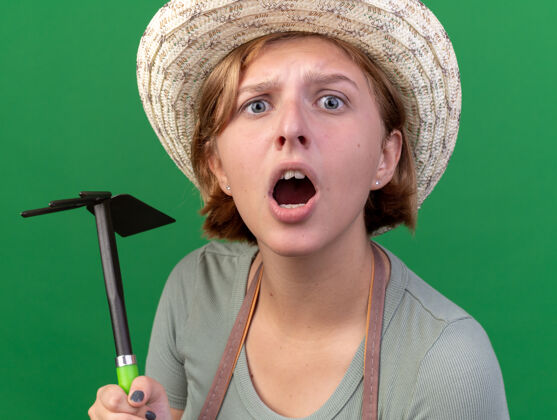 锄头不知所措的年轻斯拉夫女园丁戴着园艺帽手持锄头耙看着相机年轻绿色女性