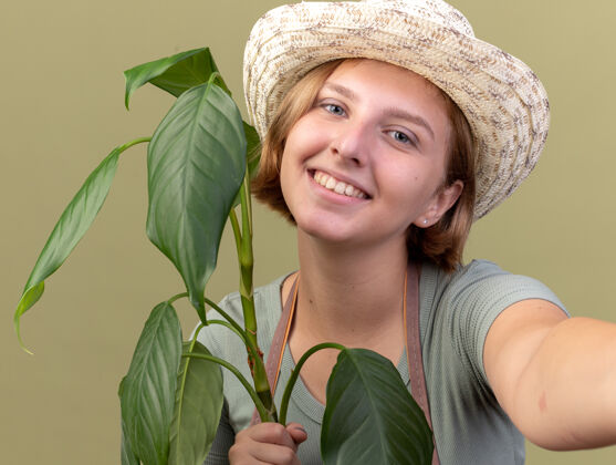橄榄微笑着的年轻斯拉夫女园丁戴着园艺帽拿着植物假装拿着相机自拍帽子采取微笑