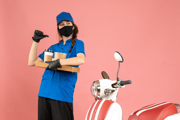 指向俯视图自信的信使女孩戴着医用口罩手套站在摩托车旁拿着咖啡小蛋糕指着背面的粉彩桃色背景面具摩托车举行