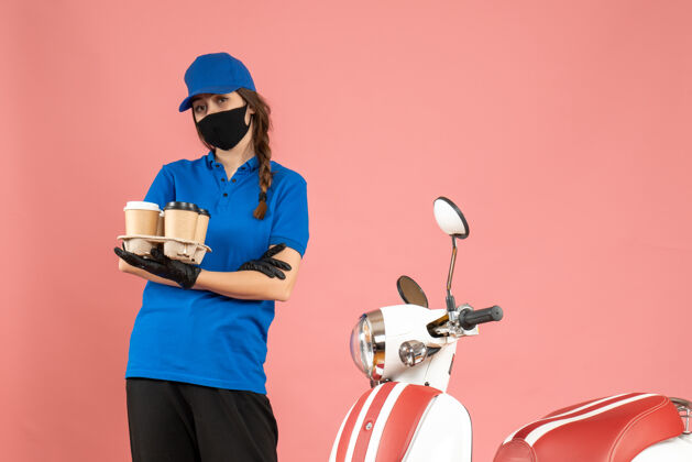 面具正面图：戴着医用口罩手套的年轻快递女孩站在摩托车旁 手里拿着粉色背景上的咖啡小蛋糕背景举行小