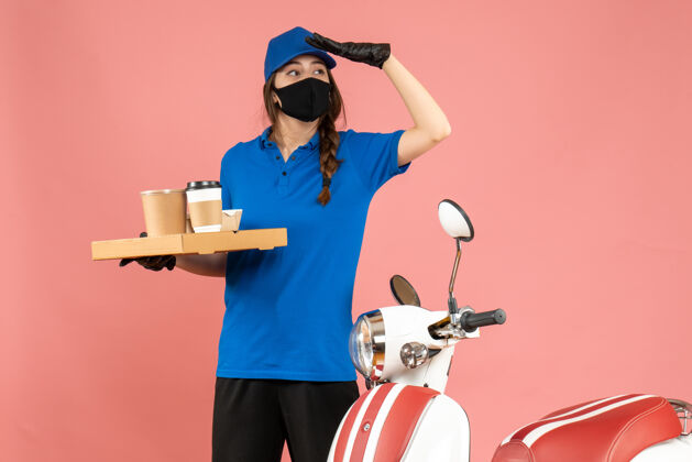 男前视图聚焦快递女孩戴着医用口罩手套站在摩托车旁边拿着咖啡小蛋糕粉彩桃红色背景人物粉彩咖啡