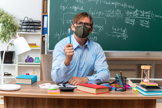 展示年轻的男老师戴着眼镜 戴着口罩 一脸严肃地拿着铅笔坐在课桌旁 教室的黑板前放着书和笔记年轻佩戴黑板