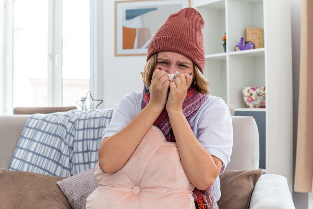 组织不健康的年轻女士戴着暖和的帽子 戴着围巾 看起来不舒服 生病 拿着纸巾 患感冒和流感 在光线充足的客厅里坐在沙发上打喷嚏不适鼻子生活