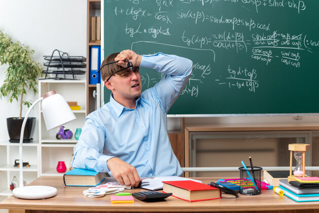 笔记戴着眼镜的年轻男老师坐在教室的黑板前 手里拿着书和笔记 神情迷茫 非常焦虑焦虑学校困惑
