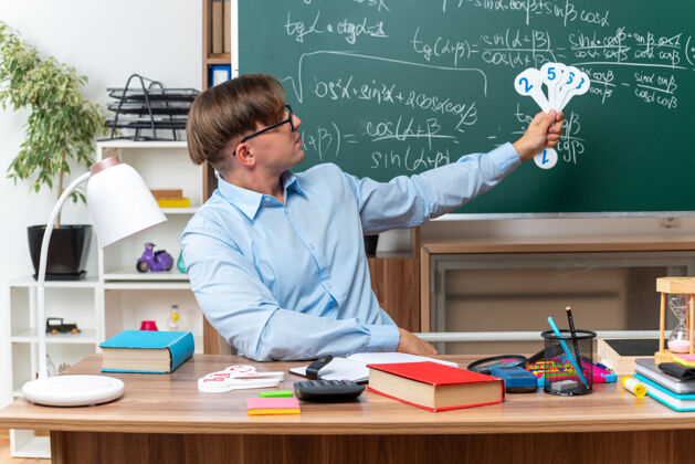 课桌年轻的男老师戴着眼镜 展示着车牌 微笑着 自信地坐在课桌旁 教室的黑板前放着书和笔记学校展示教室