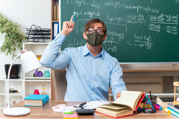 目录年轻的男老师戴着眼镜 戴着口罩 显示食指有了新的想法 坐在课桌旁 教室的黑板前放着书和笔记青少年笔记展示