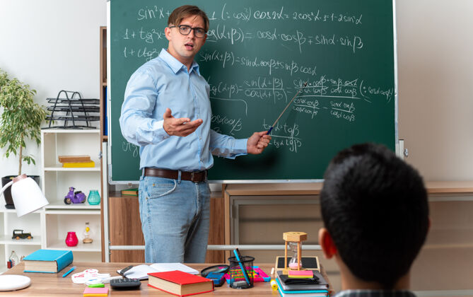 困惑戴着眼镜的年轻男教师 站在教室里数学公式的黑板旁 神情困惑 不高兴地向学生们讲解功课站黑板年轻
